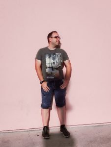 Man with glasses and a t-shirt stands against a pink wall, looking off to the side.